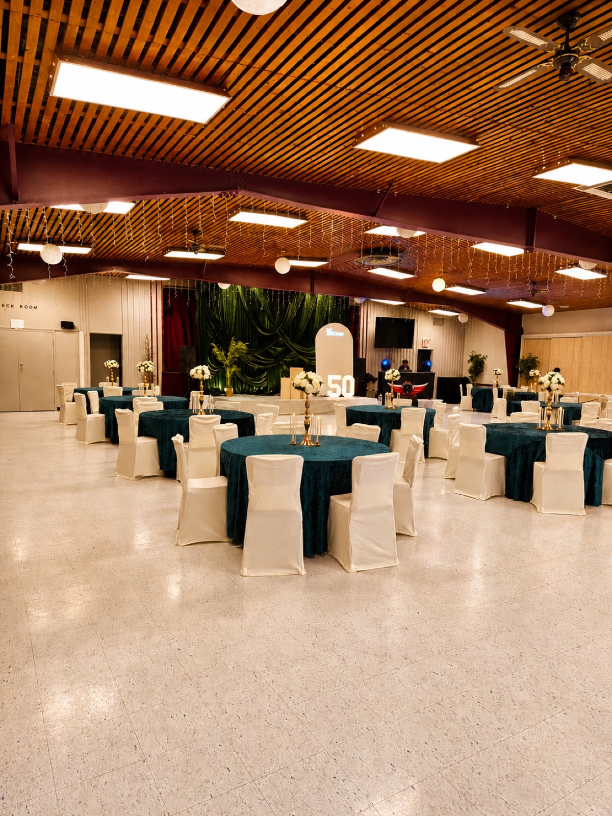 The main hall set for a celebration with round tables in teal linens, cream chair covers and a decorated stage